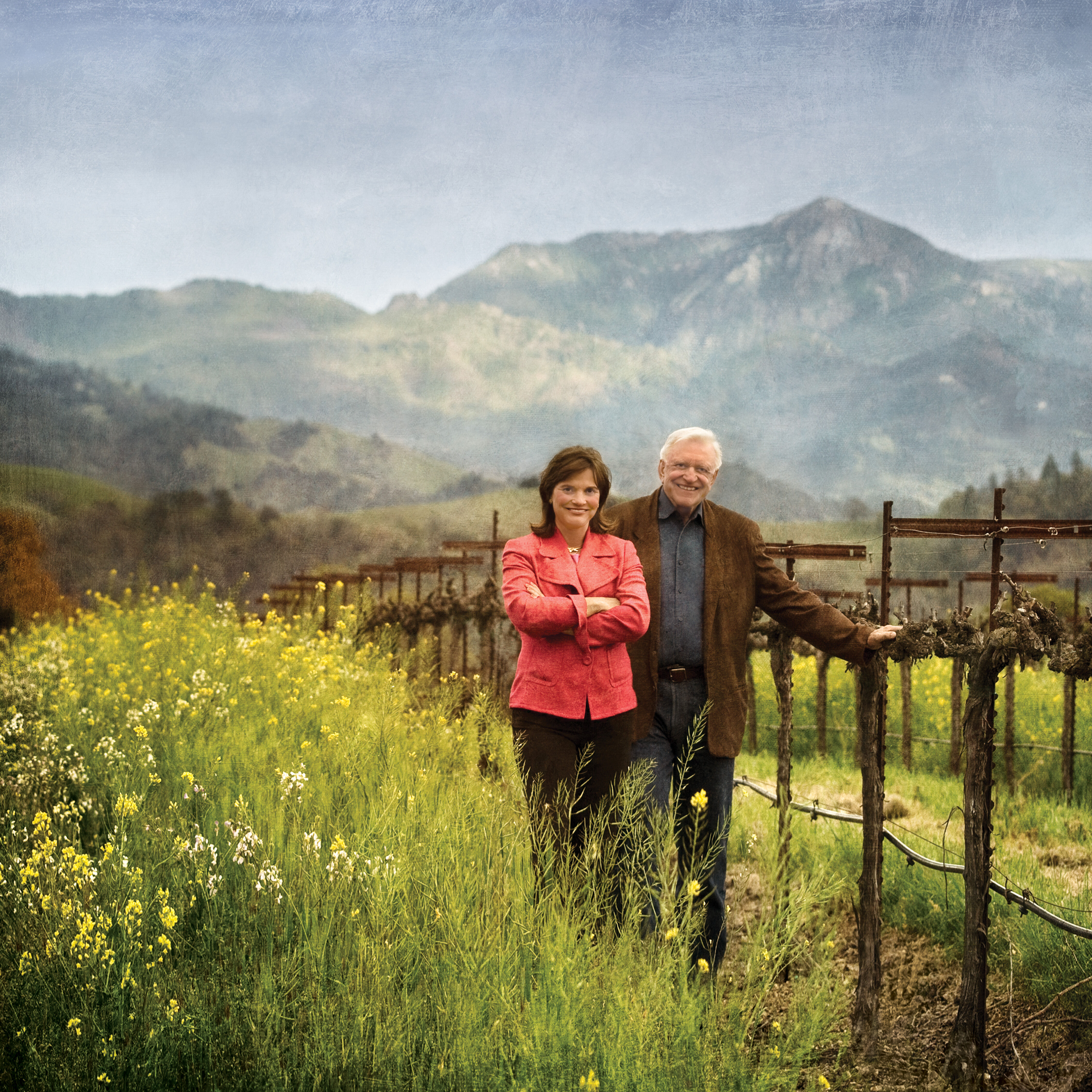 A photo of Jess and Barbara Jackson in a vineyard with mustard flowers and a mountain in the background.